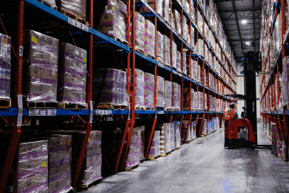 Interior view of a cold storage warehouse with high shelves packed with pallets wrapped in purple plastic, under dim lighting. A forklift is operated by a worker in the central aisle.