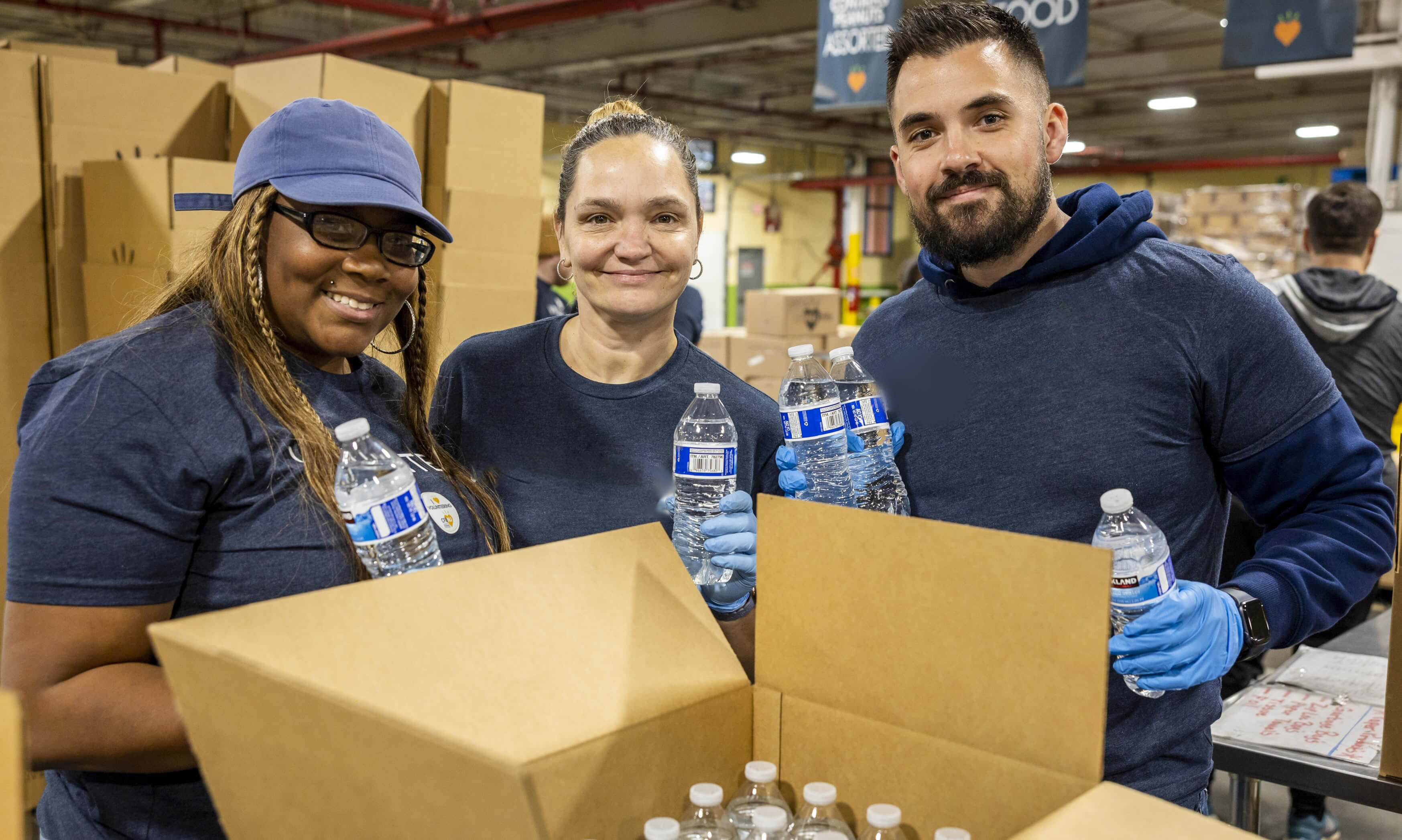 Three volunteers packing water bottles for distribution, wearing blue 'VOLUNTEER' t-shirts and smiling.